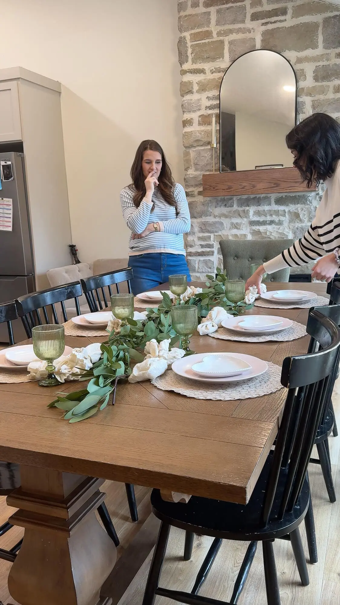 A partially set dining table featuring heart-shaped plates, green glass goblets, and eucalyptus garland. A woman adjusts a woven placemat while another stands nearby in a striped shirt, overseeing the setup.